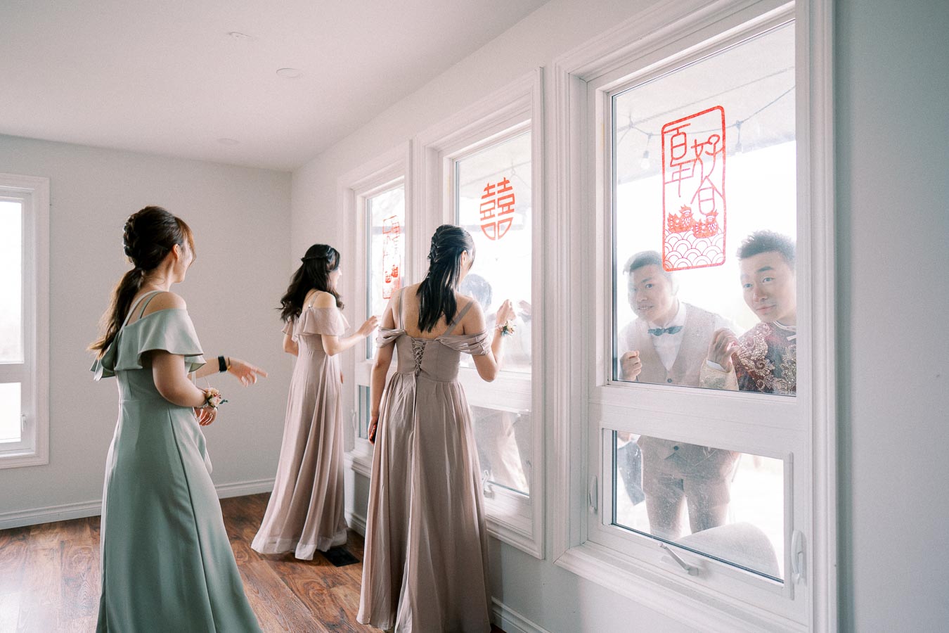 Bridesmaids wearing elegant dresses look out a window decorated with red Chinese wedding symbols as two men in suits stand outside smiling.