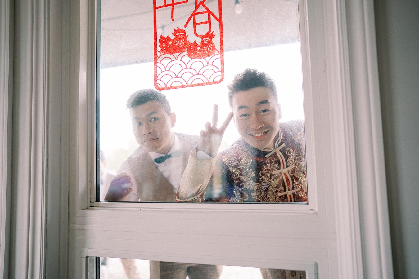 Two men smiling and looking through a window, one making a peace sign, with festive red Chinese decorations on the glass.