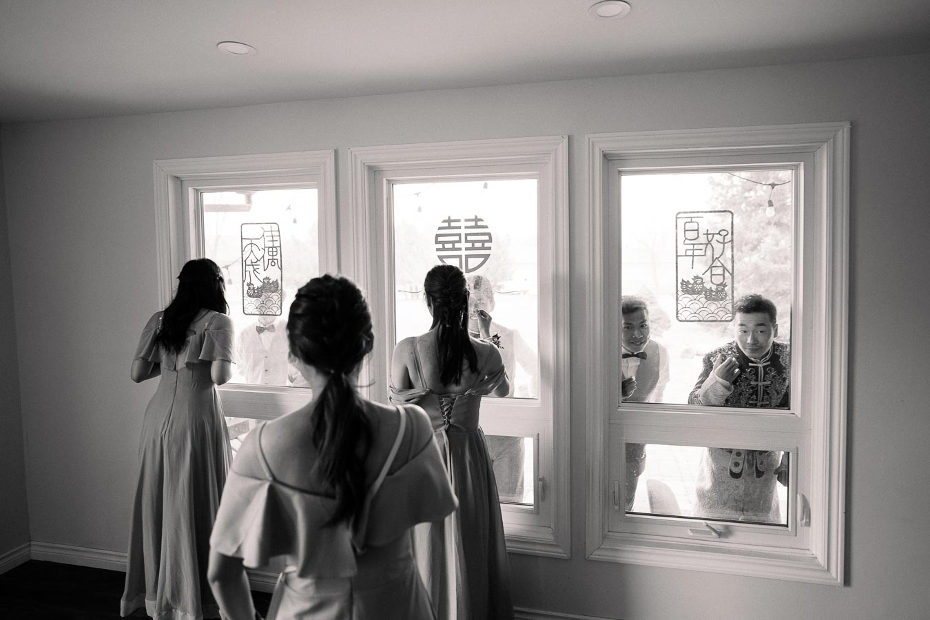 A black and white image of a bridal party inside a room, with three women facing a window. Two men peer through the window from outside. Chinese characters and traditional decorations are on the glass, creating a festive and cultural atmosphere. The image captures anticipation and joy, typical of a wedding day celebration.