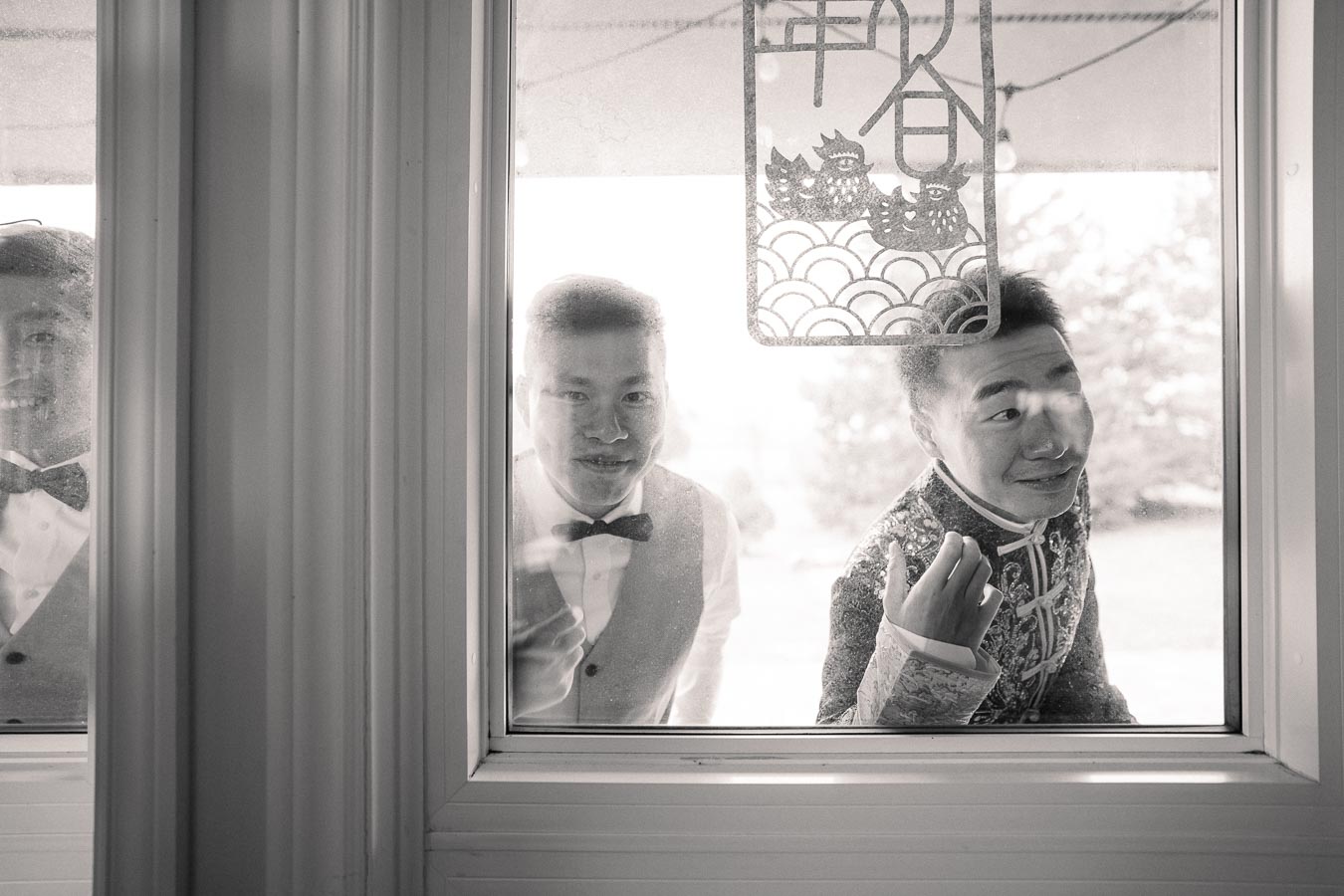 Black and white image of three men dressed in formal attire and traditional clothing looking through a window, with a Chinese paper decoration hanging in the foreground. The scene suggests a joyful event, possibly a wedding.