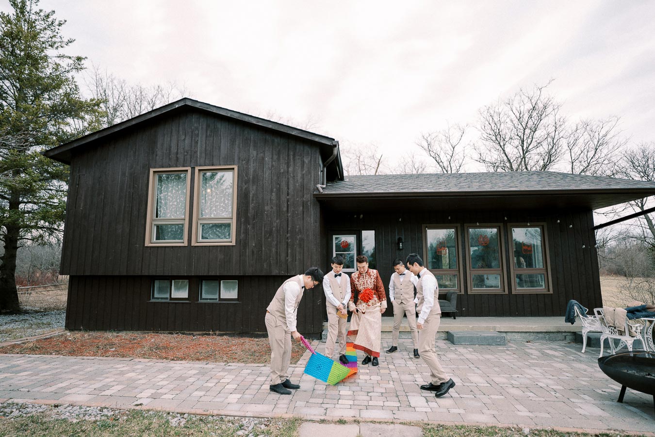 A group of people in formal attire standing on a brick patio in front of a dark wooden house, engaging with cheerful colored props. The scene presents an overcast sky and sparse trees, suggesting a casual outdoor gathering or event.