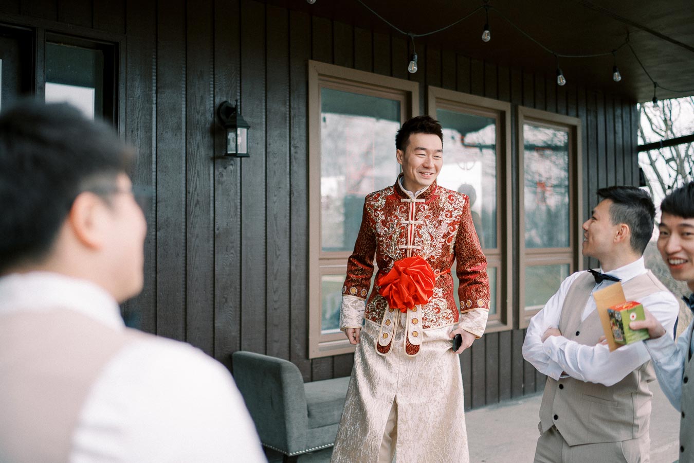 Stylishly dressed groom in traditional embroidered attire with a vibrant red pattern and decorative embellishments, standing with groomsmen who are wearing formal suits, smiling and sharing a joyous moment outdoors.
