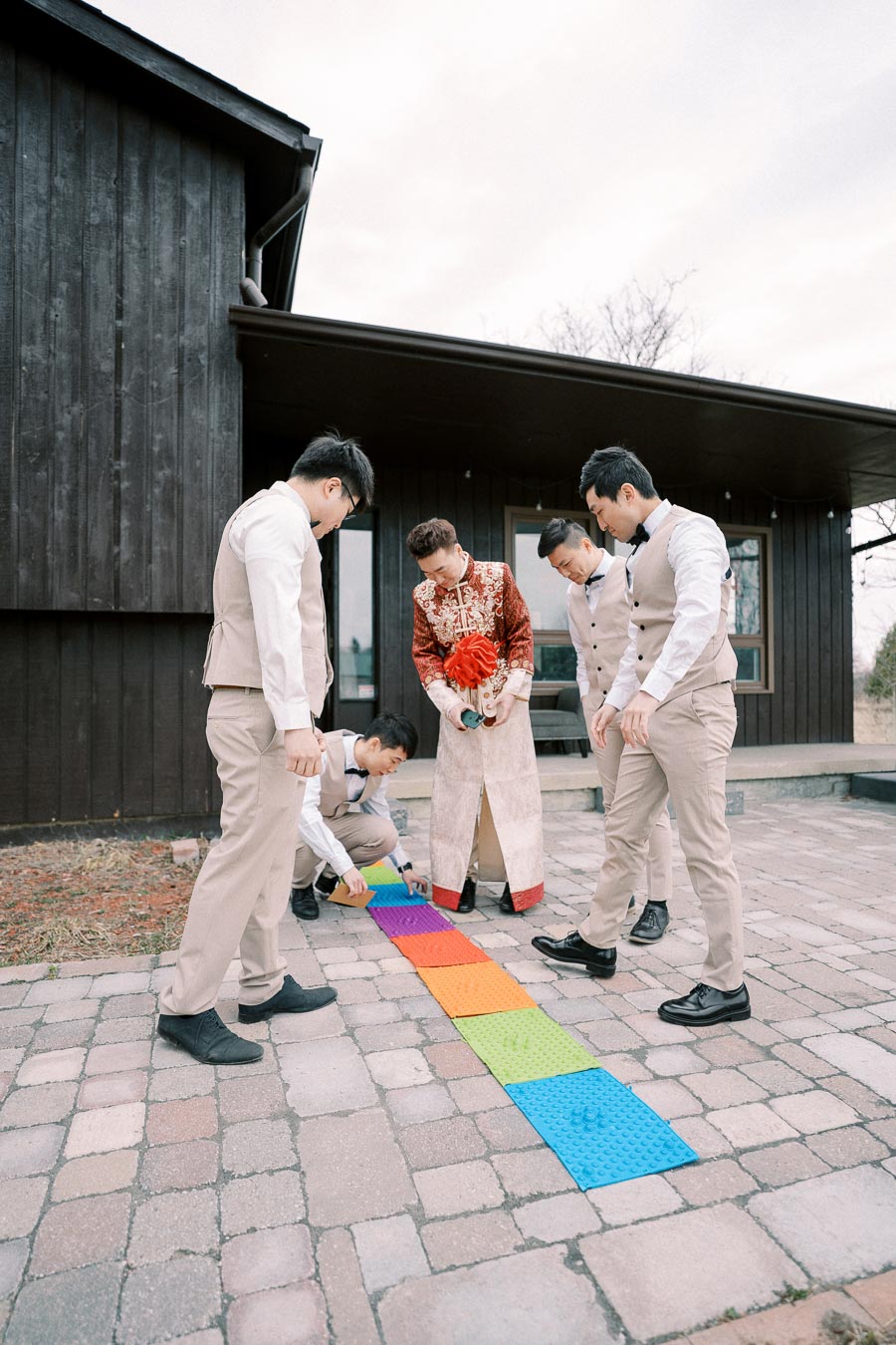 A group of men, one dressed in traditional Asian attire, engage in a playful activity involving colorful mats on a tiled patio outside a modern dark wooden building.
