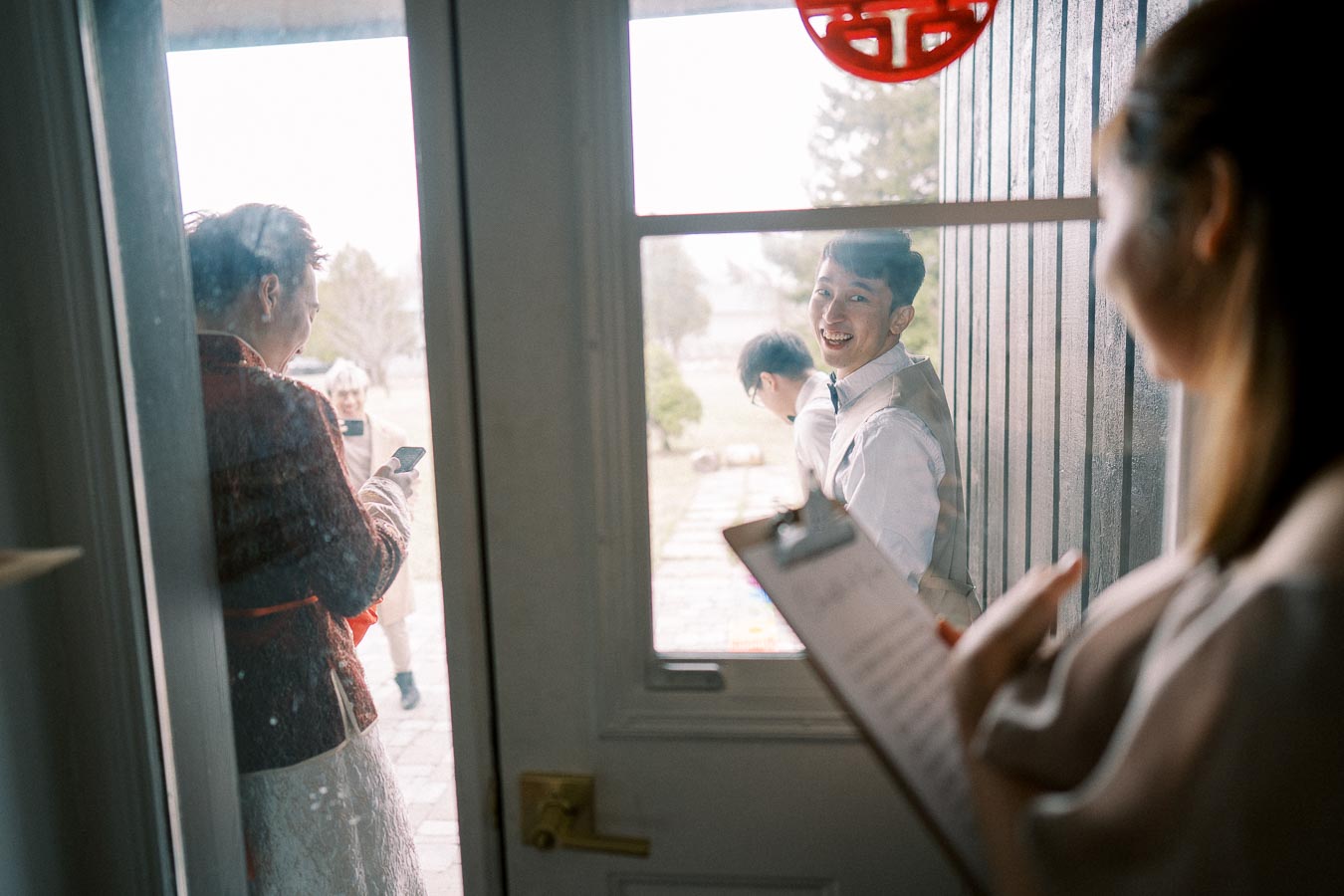A person standing outside a door smiling, as someone inside holds a clipboard. Other individuals are in the background, one using a smartphone. The scene appears to be a friendly and possibly celebratory gathering.