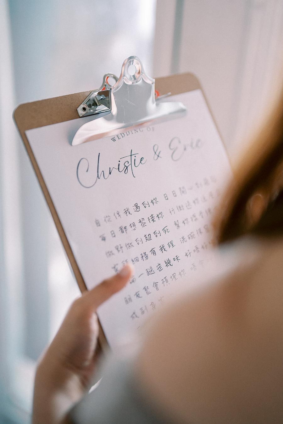 Close-up of a wedding invitation being held on a clipboard, featuring elegant handwriting and text in both English and Chinese characters, with the names 'Christie & Eric' prominently displayed.