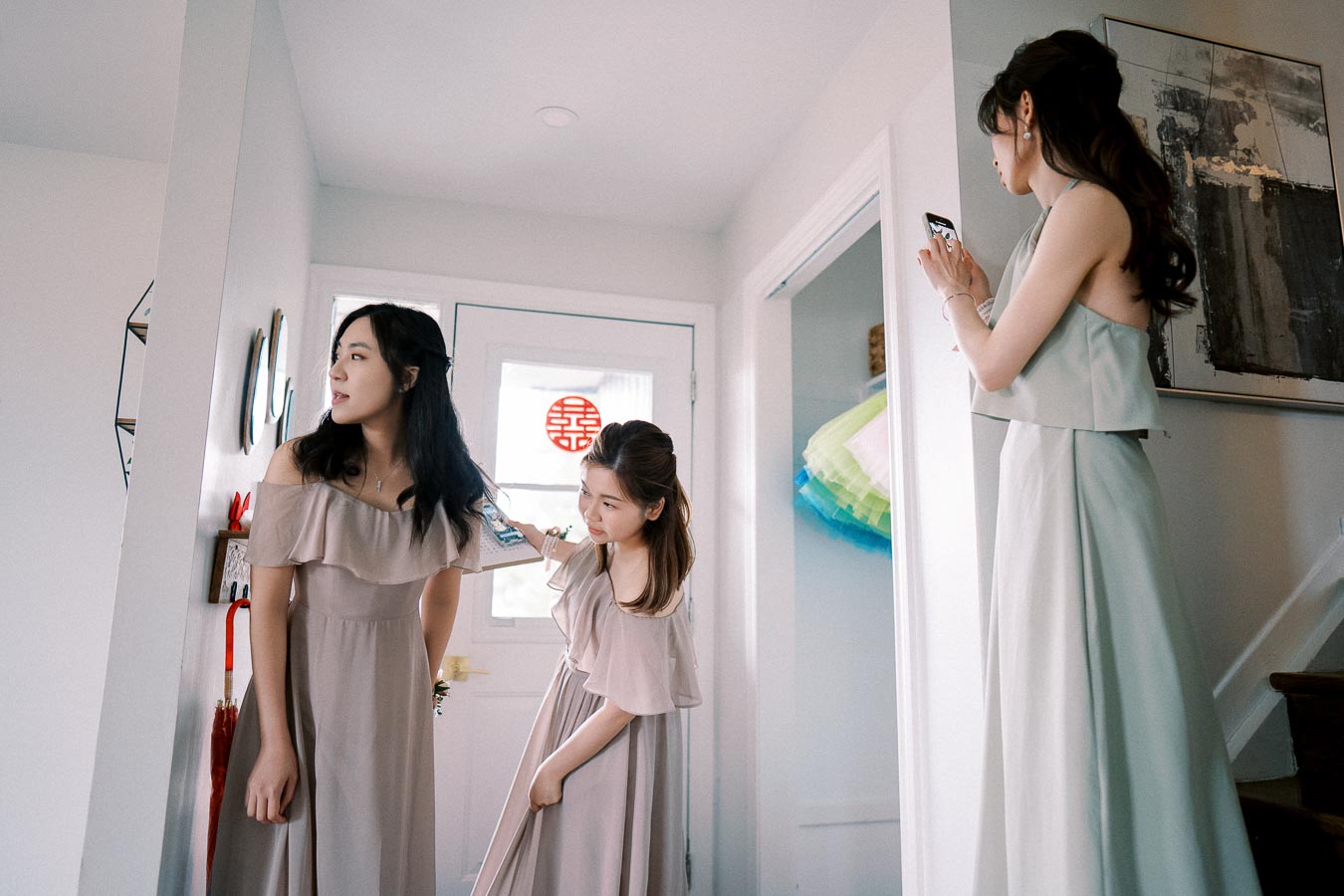 Three women in elegant dresses are preparing for a special occasion. One woman is fixing another’s hair, while the third is taking a photo with a smartphone. The setting appears to be a bright, modern home interior, with decorative elements visible in the background.