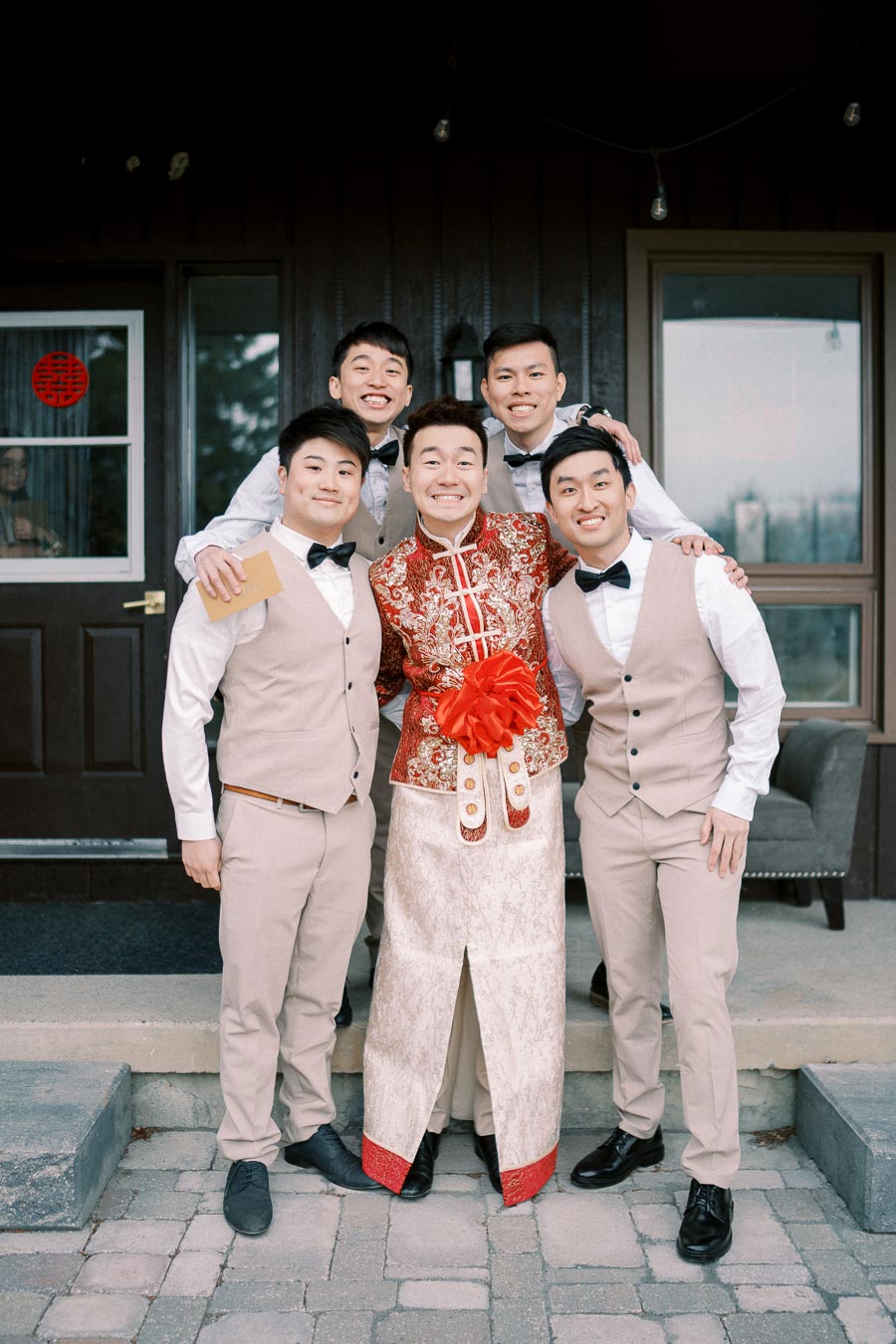 A group of five men posing in front of a house, with one individual wearing a traditional Chinese wedding outfit and the others dressed in tan vests and bow ties, celebrating a wedding occasion.