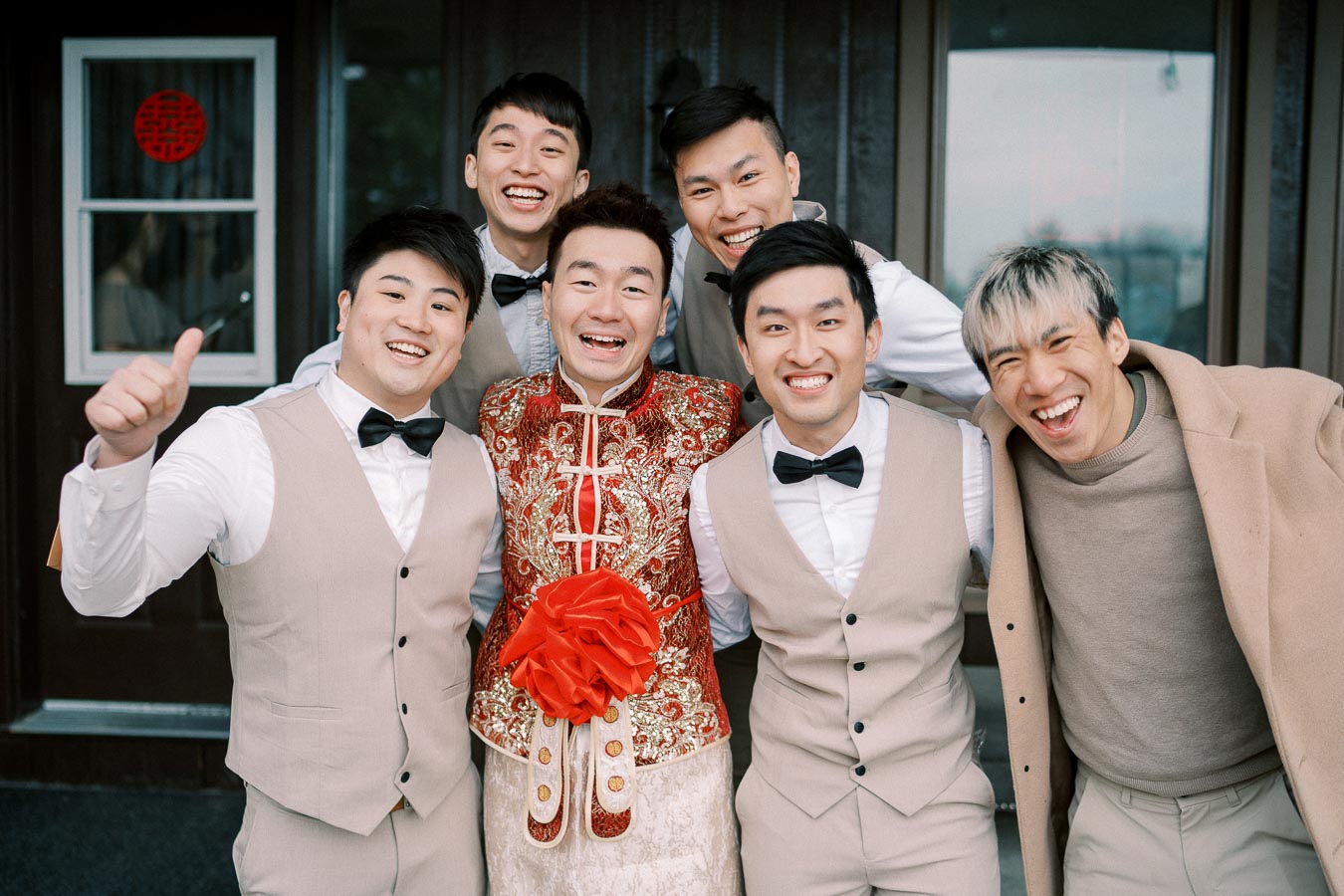 A happy groom in traditional red wedding attire poses with groomsmen in matching vests and bow ties.