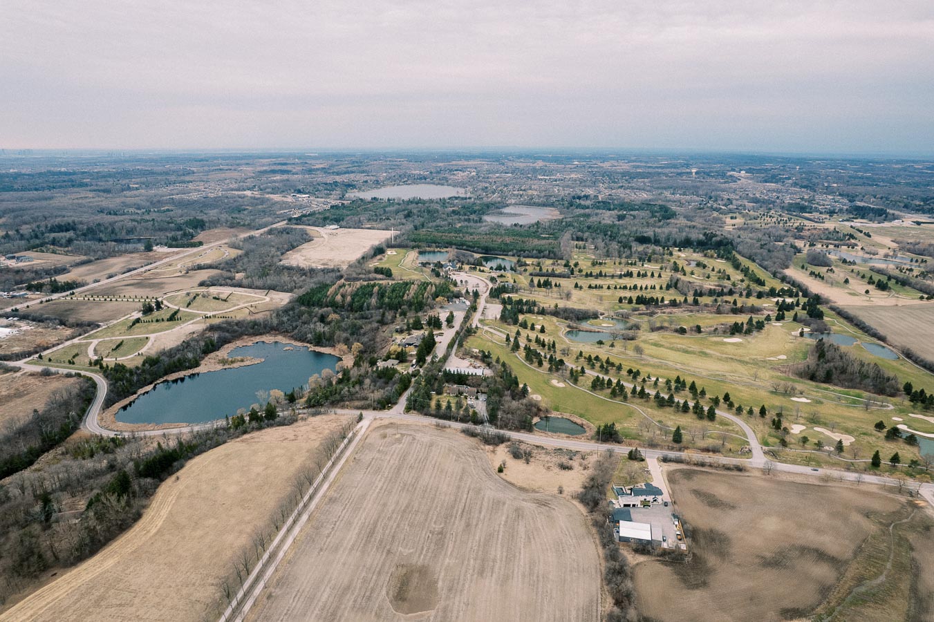 Aerial view of a sprawling rural landscape featuring a golf course with green fairways and small water bodies, surrounded by fields and woodlands under a cloudy sky.
