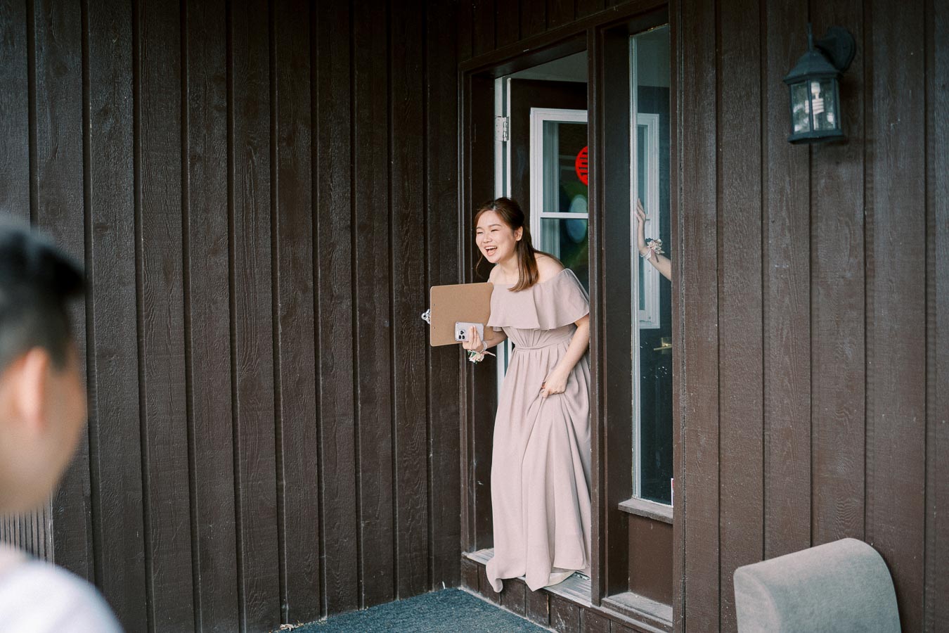 A woman in an elegant pink dress stepping outside through a doorway, holding a smartphone and smiling, against a wooden wall background.