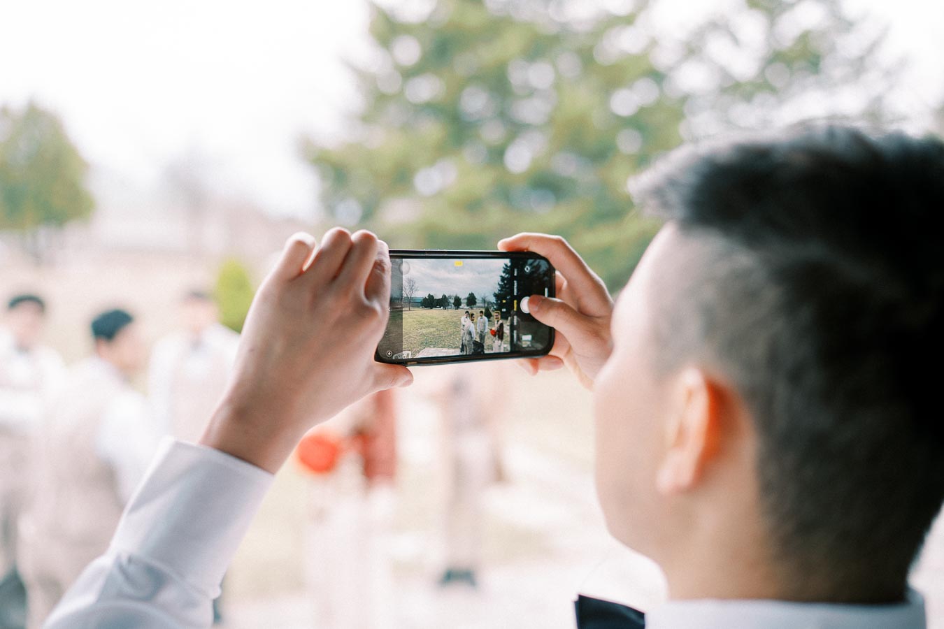 A person taking a photo of a wedding party outdoors using a smartphone, capturing a group of people dressed in formal attire, with blurred greenery in the background.