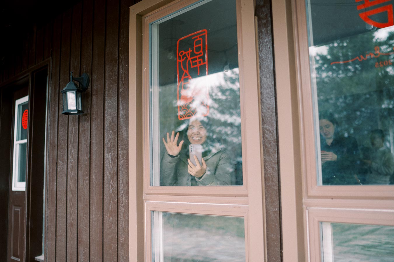 A person smiling and waving from behind a window, holding a smartphone, with a red decorative element on the glass. The scene includes reflections of trees and another person in the background, enhancing the inviting atmosphere.