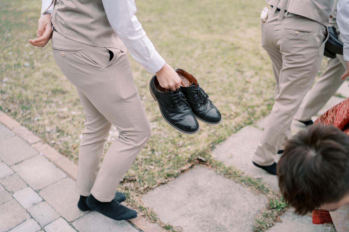 Person in beige suit holding black dress shoes while walking on a stone path outside, displaying formal attire and footwear on a grassy background.