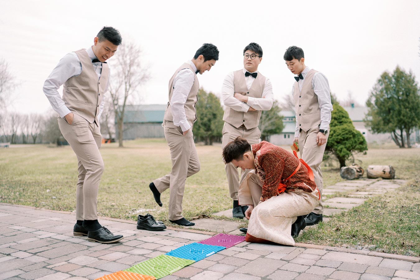 Groom kneeling on colorful textured mat during outdoor wedding ceremony, surrounded by groomsmen in matching beige suits and bow ties.