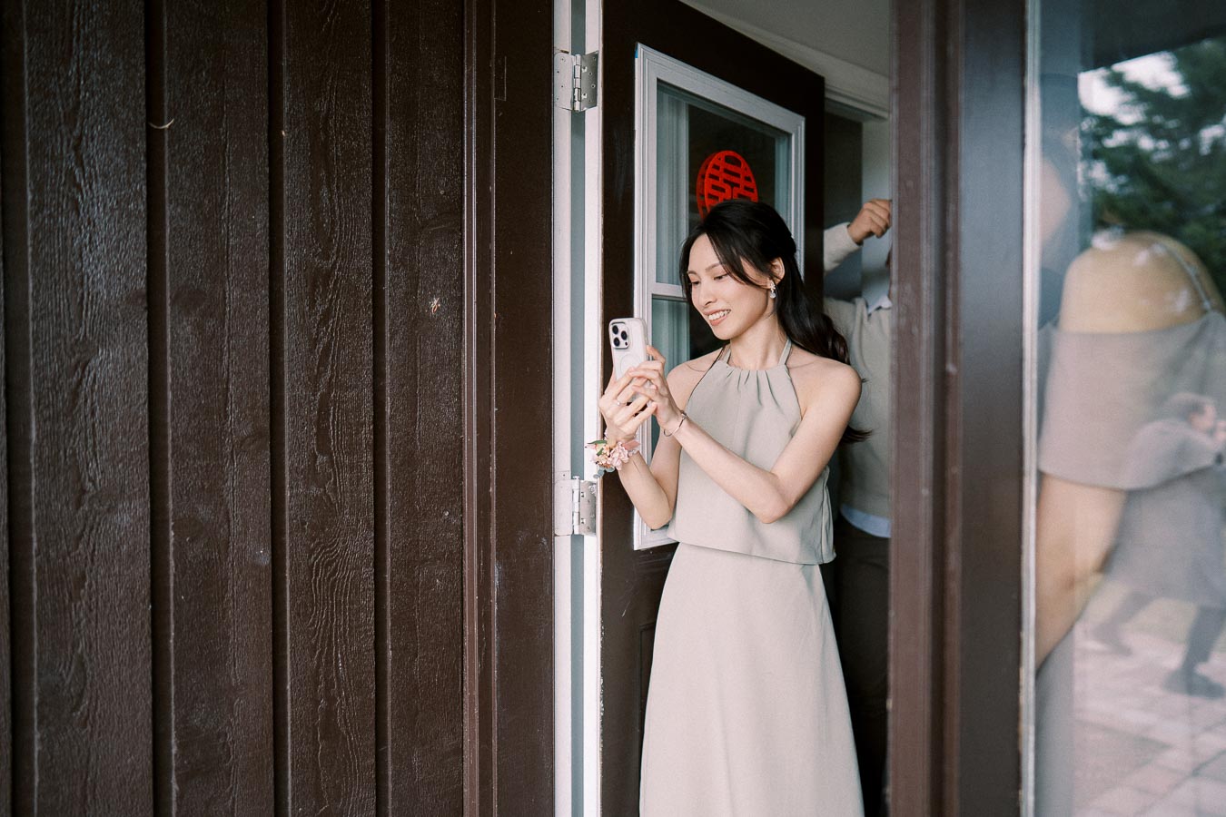 A woman in a light dress stands in an open doorway, smiling while looking at her smartphone, with a wooden wall in the background.