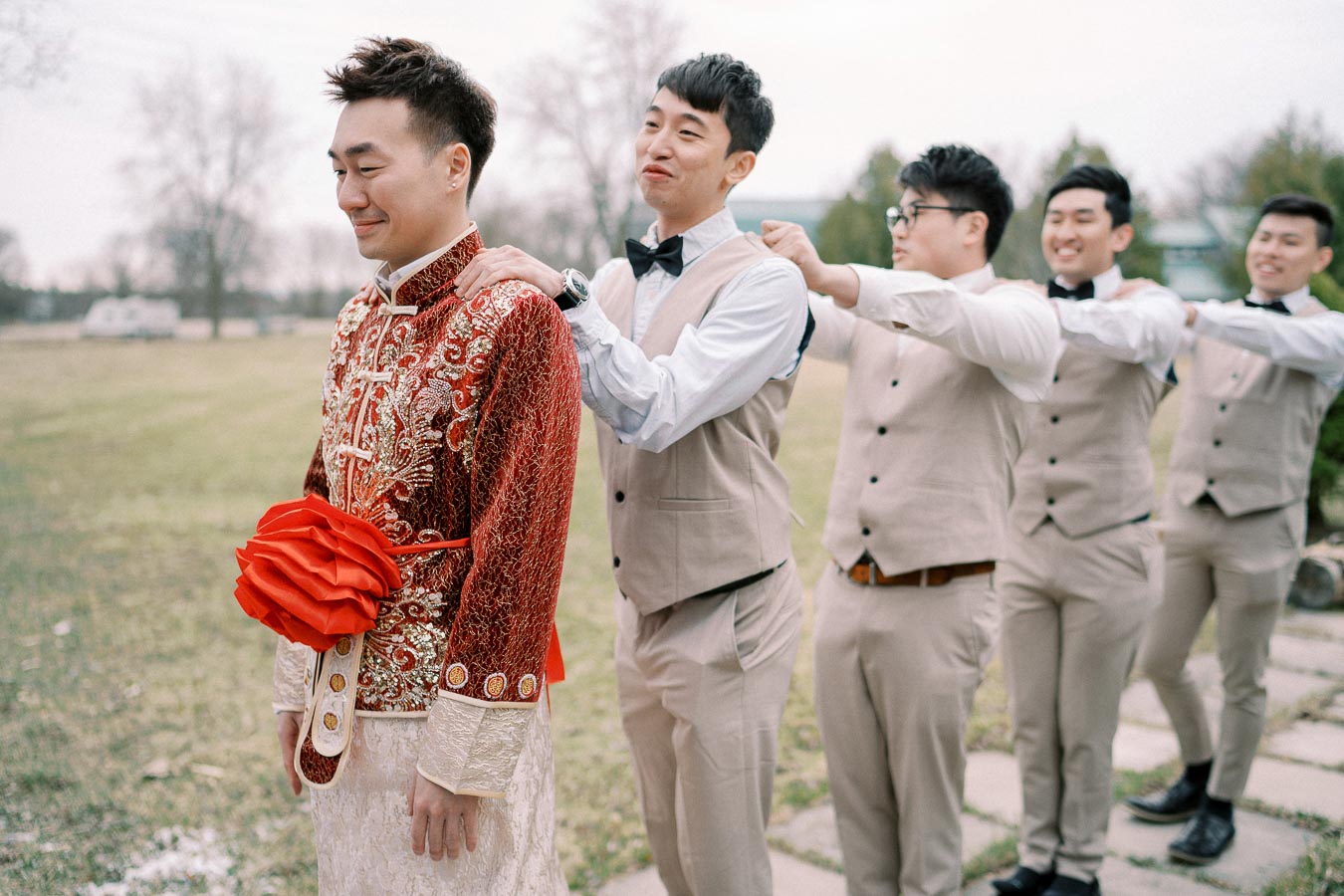 A groom in traditional Chinese wedding attire standing in a line outdoors with groomsmen dressed in beige suits, smiling and holding onto each other's shoulders.