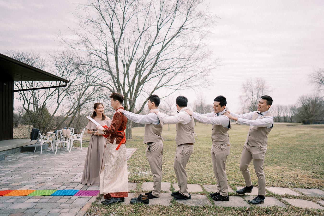 A group of men in matching beige vests and trousers play a fun outdoor wedding game, lined up behind the groom dressed in a traditional red and gold robe, as they approach a bridesmaid holding a script on a stone path with a colorful mat, set against a backdrop of bare trees and overcast sky.