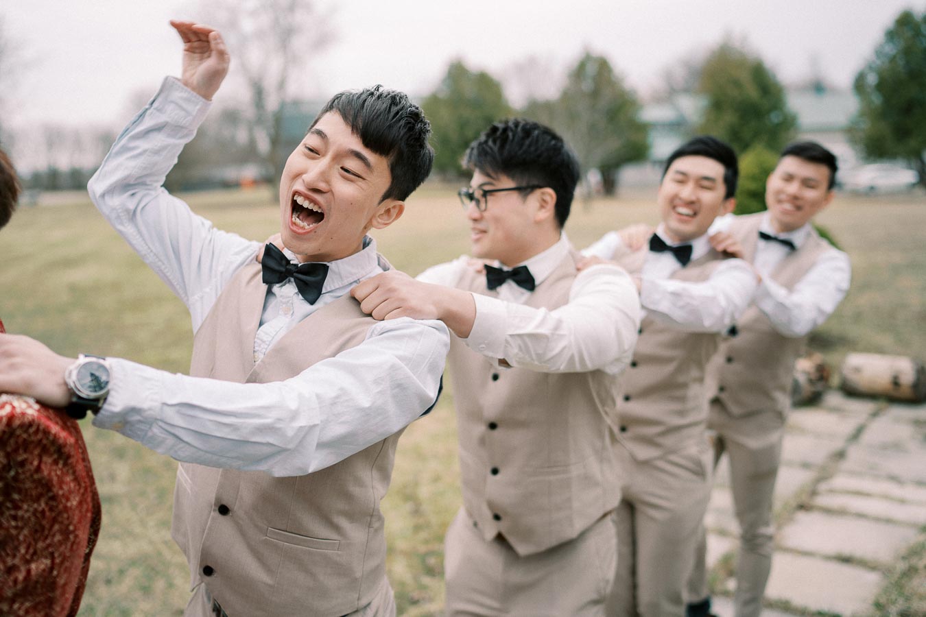 A group of groomsmen in matching beige vests and black bow ties joyfully dancing in a line outdoors, showcasing a fun and playful atmosphere at a wedding celebration.