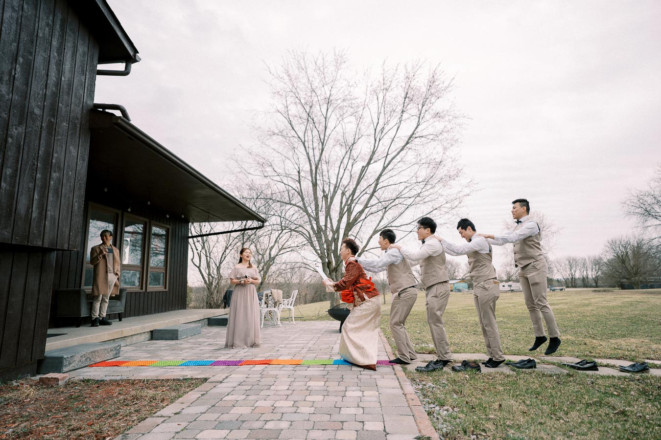 Wedding party playing a humorous game outside a rustic house, featuring a rainbow-colored mat, groomsmen in beige suits and bow ties hopping behind the bride in a traditional gown, with a bridesmaid and speaker watching nearby.