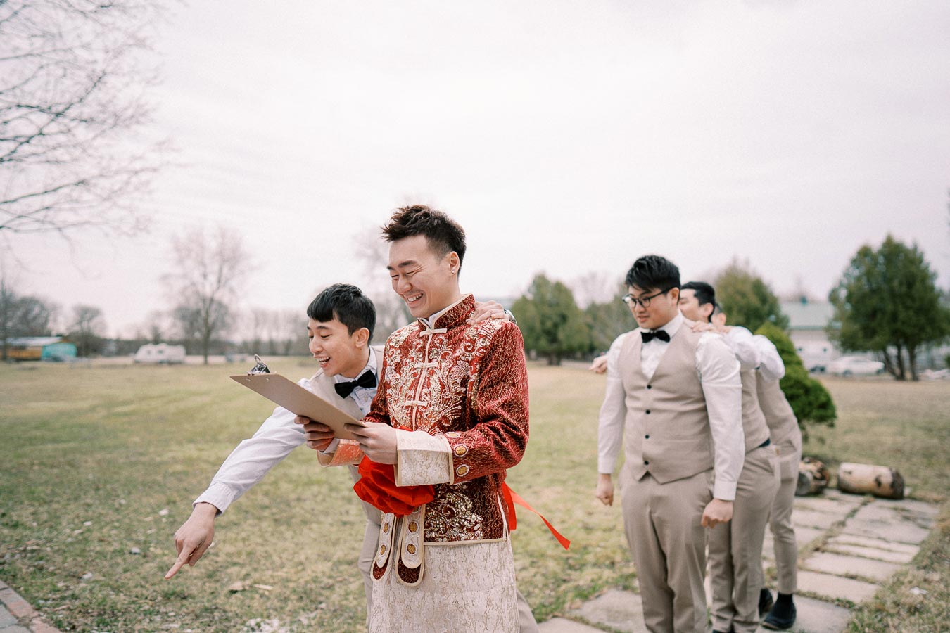 Groom in a traditional embroidered outfit, laughing with groomsmen in tan suits and bow ties, outdoors on a grassy field with trees in the background.