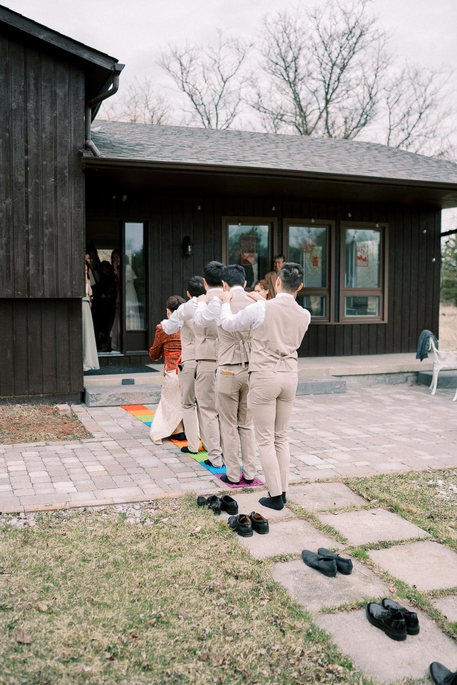 A group of people in matching outfits, participating in an outdoor event, walk in a line on a multicolored mat leading to a wooden building, with shoes removed and left on the grass beside the walkway.