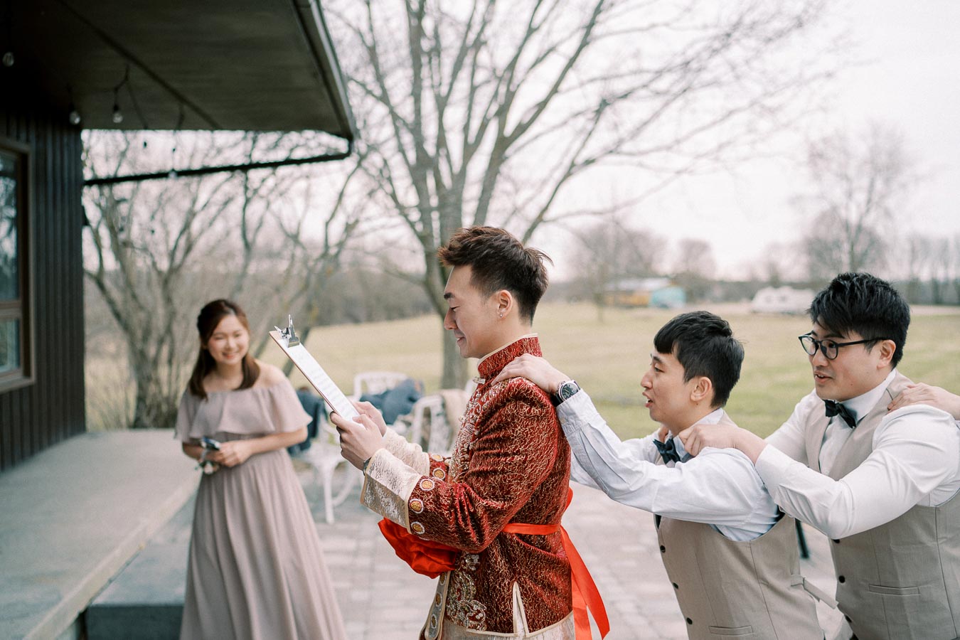 A group of friends having fun at an outdoor wedding, with a smiling groom in a traditional red embroidered outfit holding a clipboard, accompanied by groomsmen in light vests. A bridesmaid in a long, off-shoulder dress stands nearby, all set against a scenic, natural backdrop.