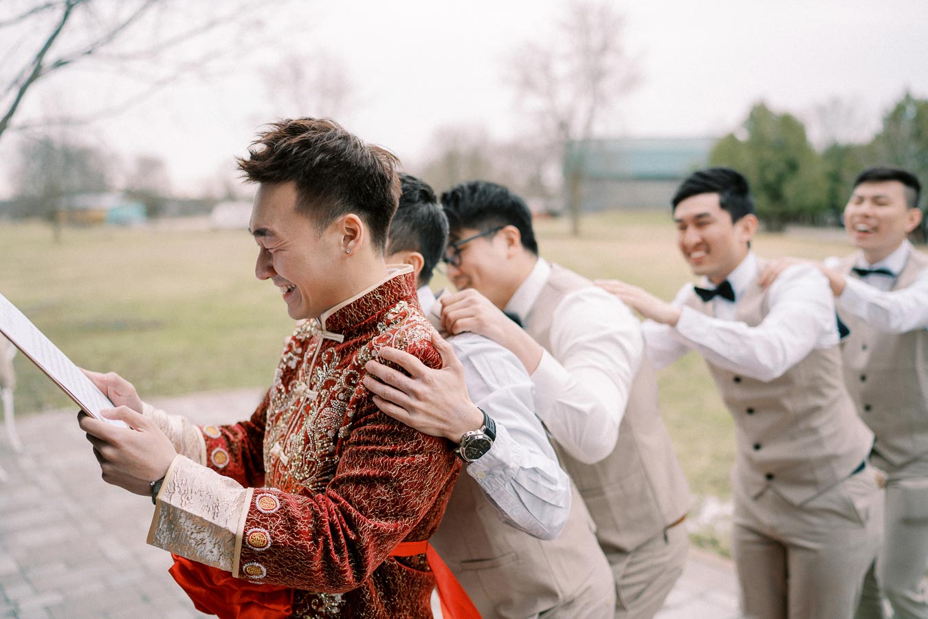 Groom in traditional red and gold embroidered attire reading a letter, with groomsmen in beige suits and bow ties playfully lined up behind him. Outdoor wedding setting with greenery in the background.