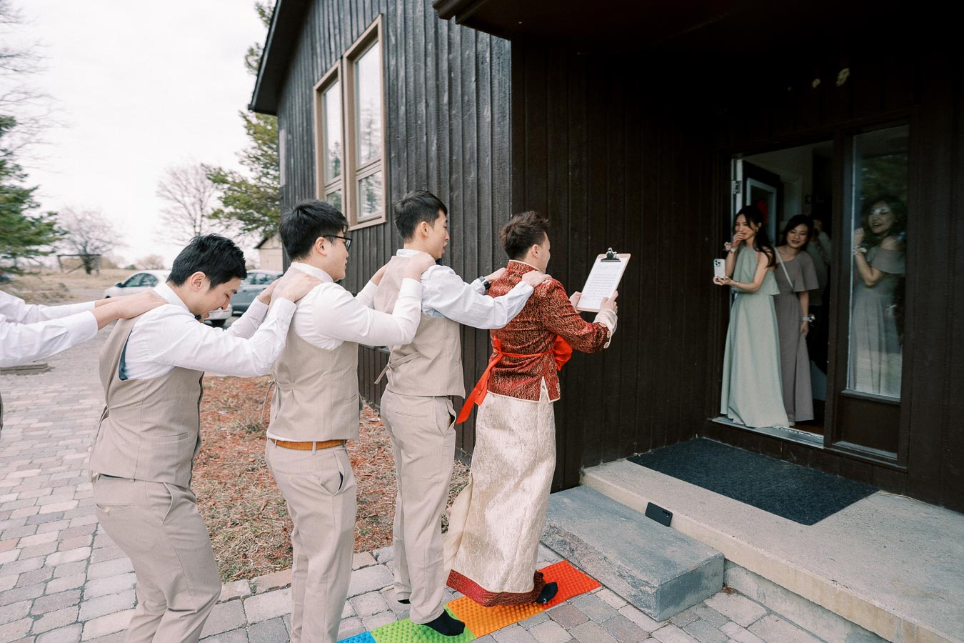 A group of men in beige suits follow a person in a traditional red and gold outfit holding a clipboard, approaching a door where women in light dresses wait, creating a festive and celebratory atmosphere.