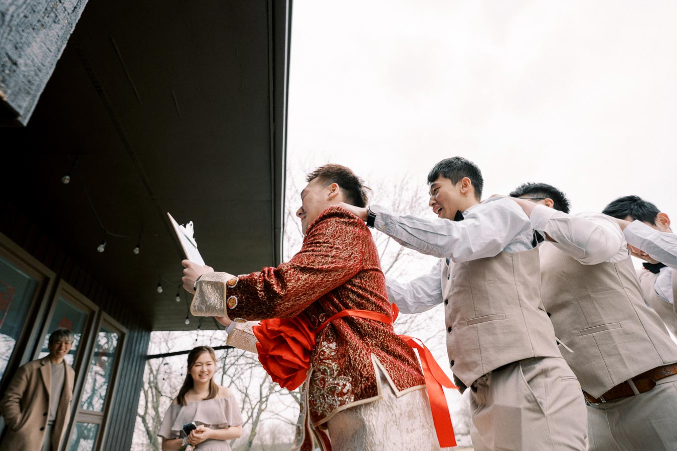 A group of groomsmen in coordinated beige suits follow a groom in a traditional ornate red and gold wedding outfit during a lively celebration. The joyful scene is set outdoors, with onlookers smiling nearby, creating a festive wedding atmosphere.