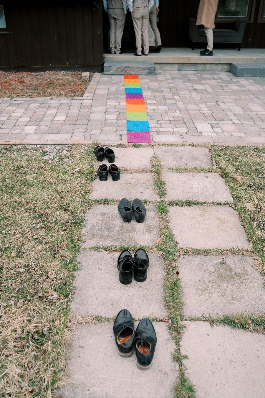 A row of black shoes placed on a stone path leading to an entrance, with a colorful mat and people standing at the doorway.
