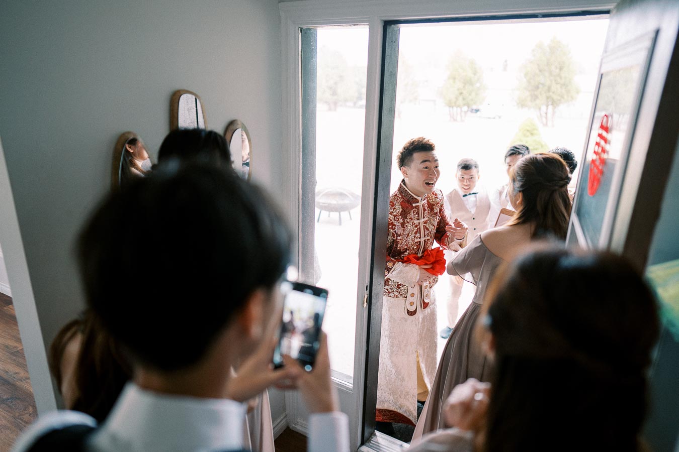 A groom in traditional attire joyfully enters a home during a wedding ceremony, surrounded by friends and family capturing the moment with their phones.