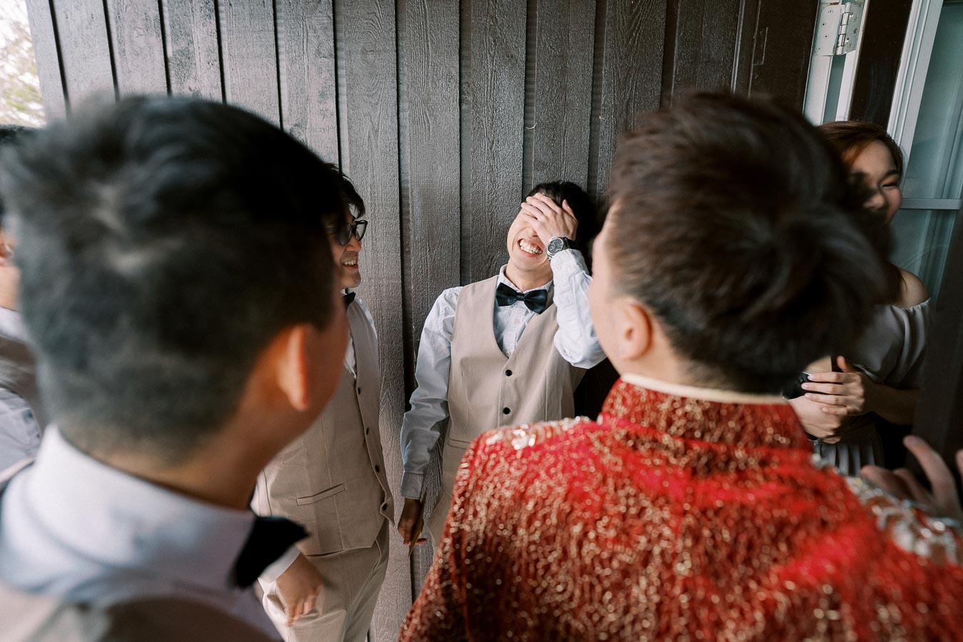 A group of people enjoying a lighthearted moment at a social gathering; one man in a vest and bow tie is playfully covering his face while laughing.