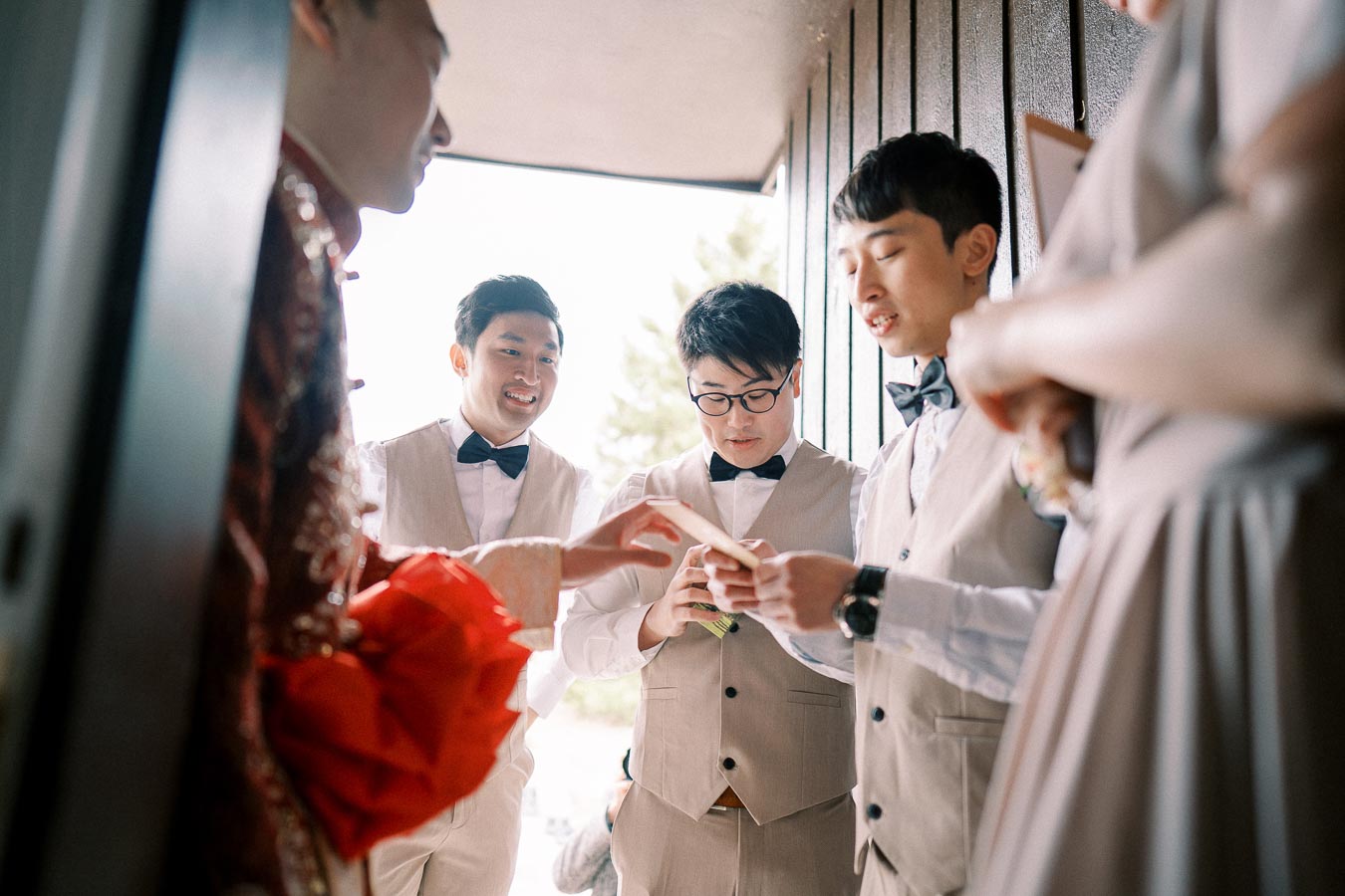 Groomsmen gathered at a wedding, wearing beige suits with bow ties, while interacting and sharing a light moment before the ceremony.