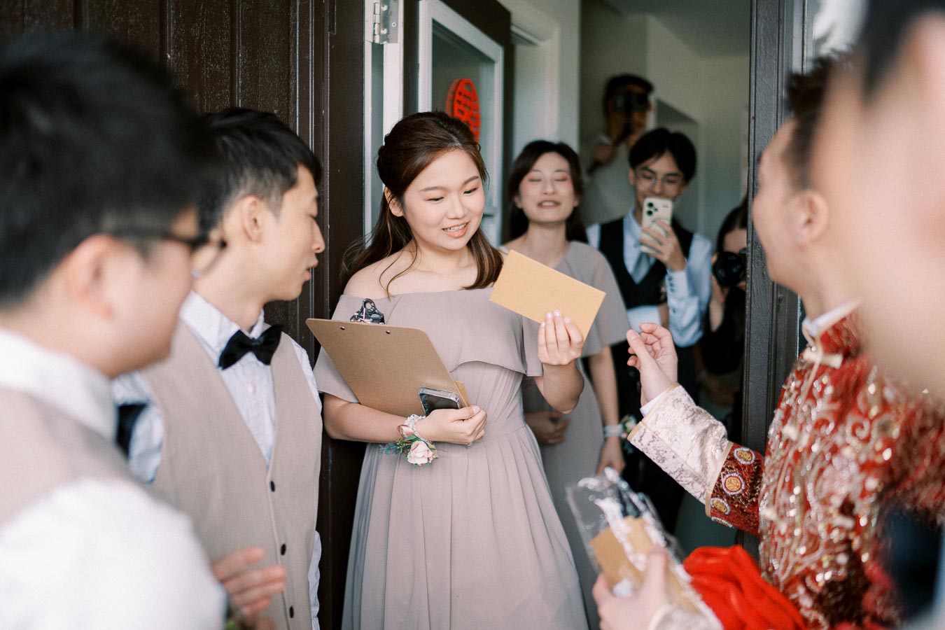 A bride and groom in traditional attire greet bridesmaids and groomsmen holding clipboards and envelopes during a wedding ceremony.