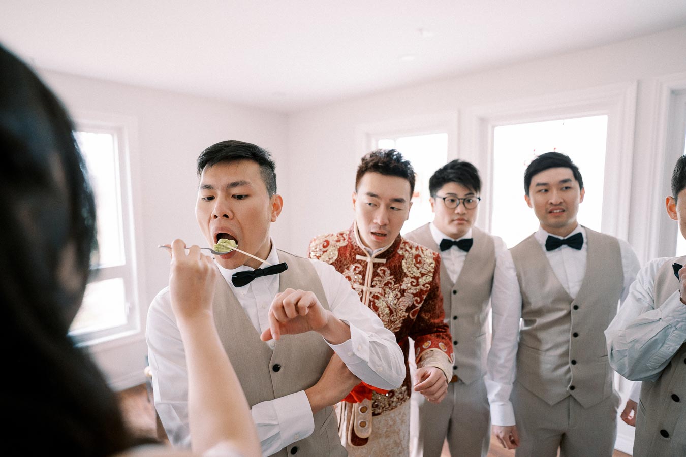 Groom and groomsmen in formal attire enjoying a playful bridal shower game with a spoon in a bright room.