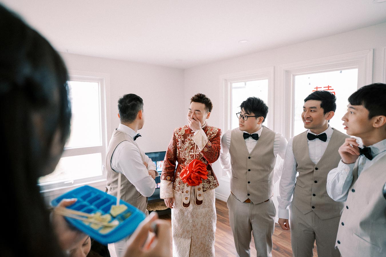 Groom surrounded by groomsmen in traditional attire, laughing and enjoying a light-hearted moment before the wedding ceremony inside a bright room.