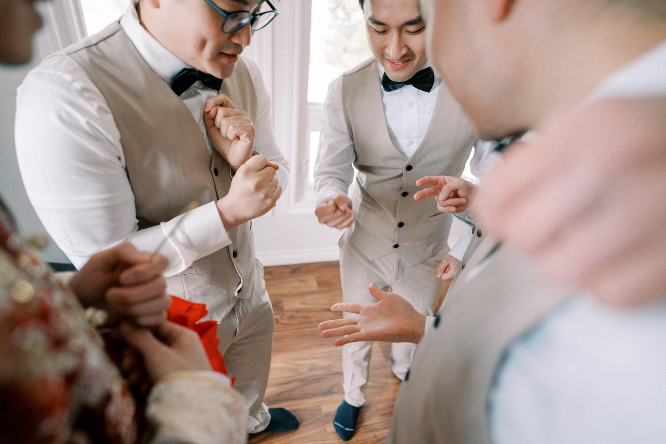 Groomsmen in beige vests play rock-paper-scissors indoors, preparing for a wedding ceremony, showcasing camaraderie and fun before the event.
