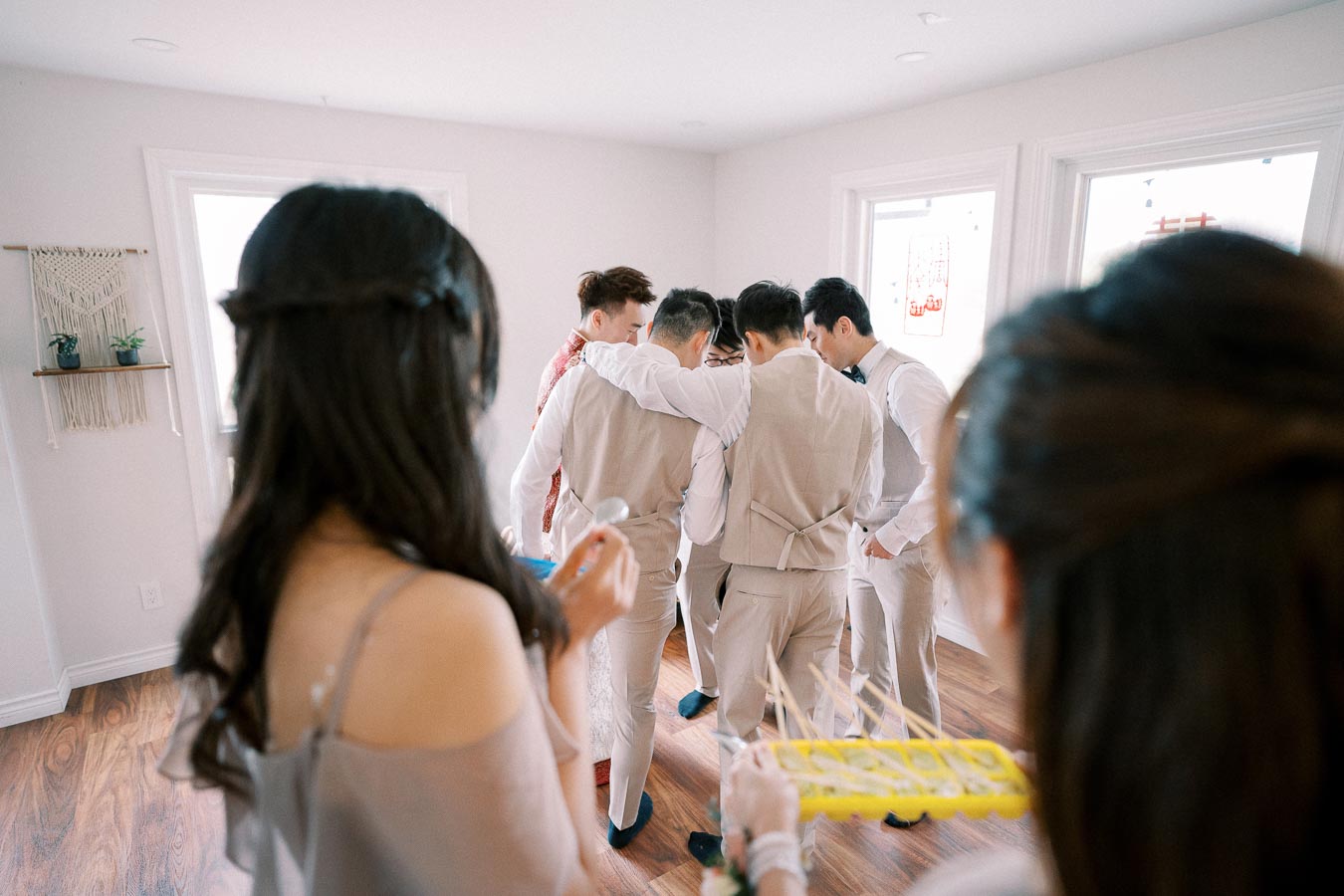 A group of groomsmen in beige suits congregate in a bright room with wooden flooring, framed by a woman in an off-shoulder dress holding a tray in the foreground, creating a candid, joyful moment before a wedding ceremony.
