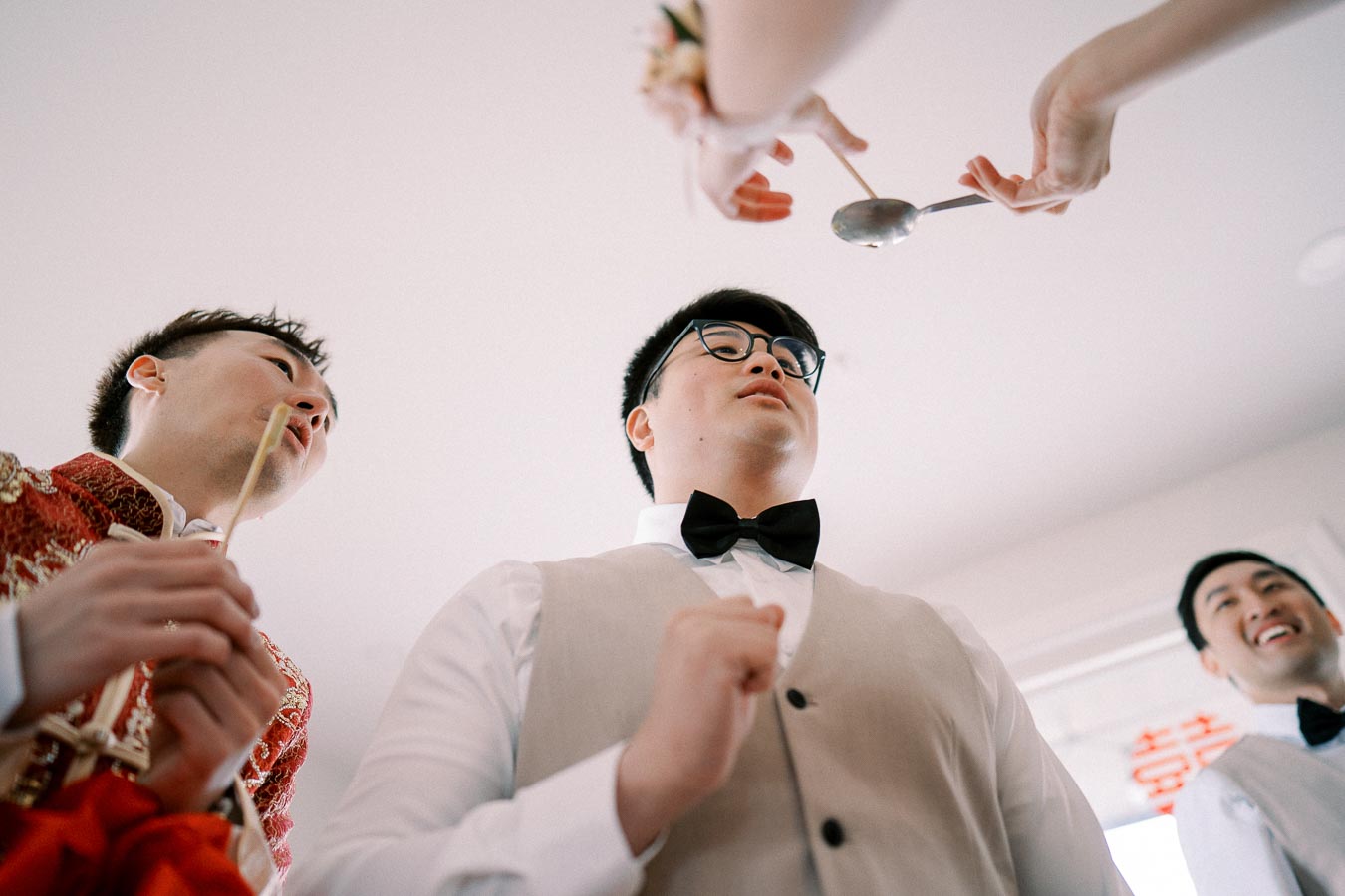 A group of three men in formal attire participating in a wedding game, with one man holding a spoon and another in traditional attire, viewed from a low angle.