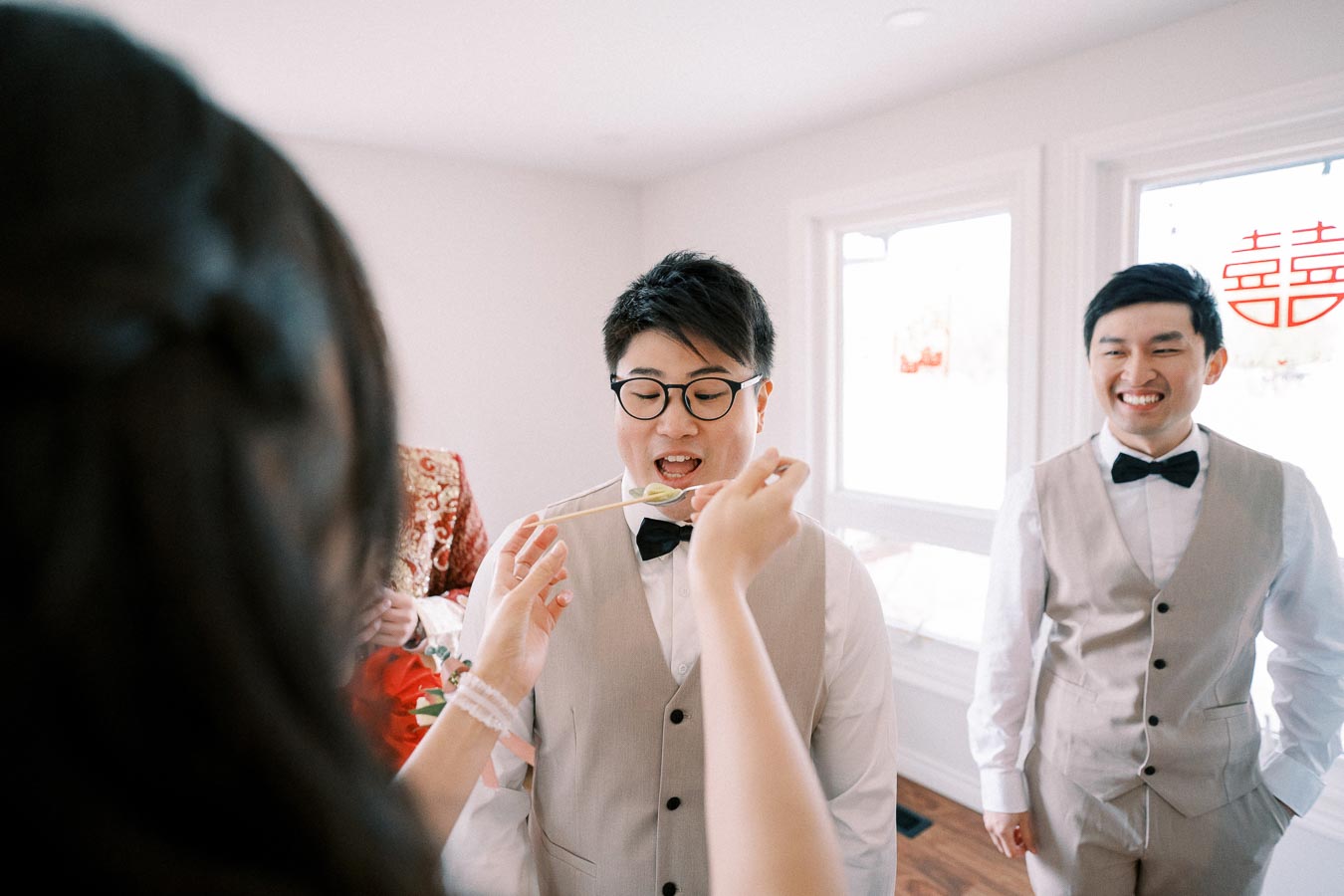 A groom in a beige suit and bow tie participating in a traditional wedding game indoors, smiling while a person feeds him with chopsticks, accompanied by another groomsman in matching attire.