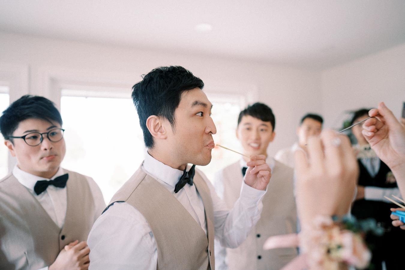 Wedding party playing fun game with chopsticks, dressed in bow ties and vests, creating a joyful atmosphere indoors.