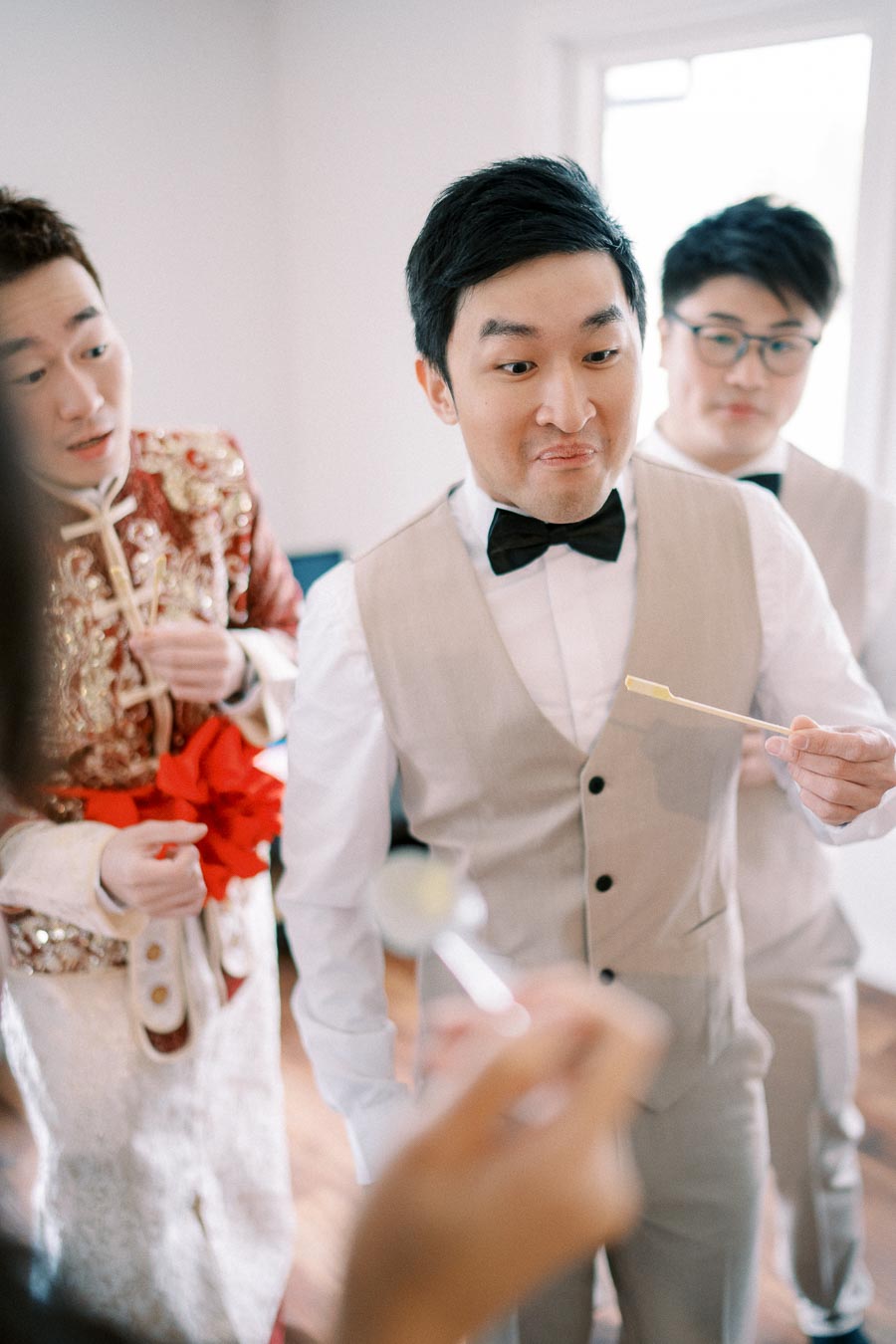 A groom and groomsmen in suits gather before a wedding ceremony, displaying playful expressions and gestures, creating a lighthearted pre-wedding moment indoors.