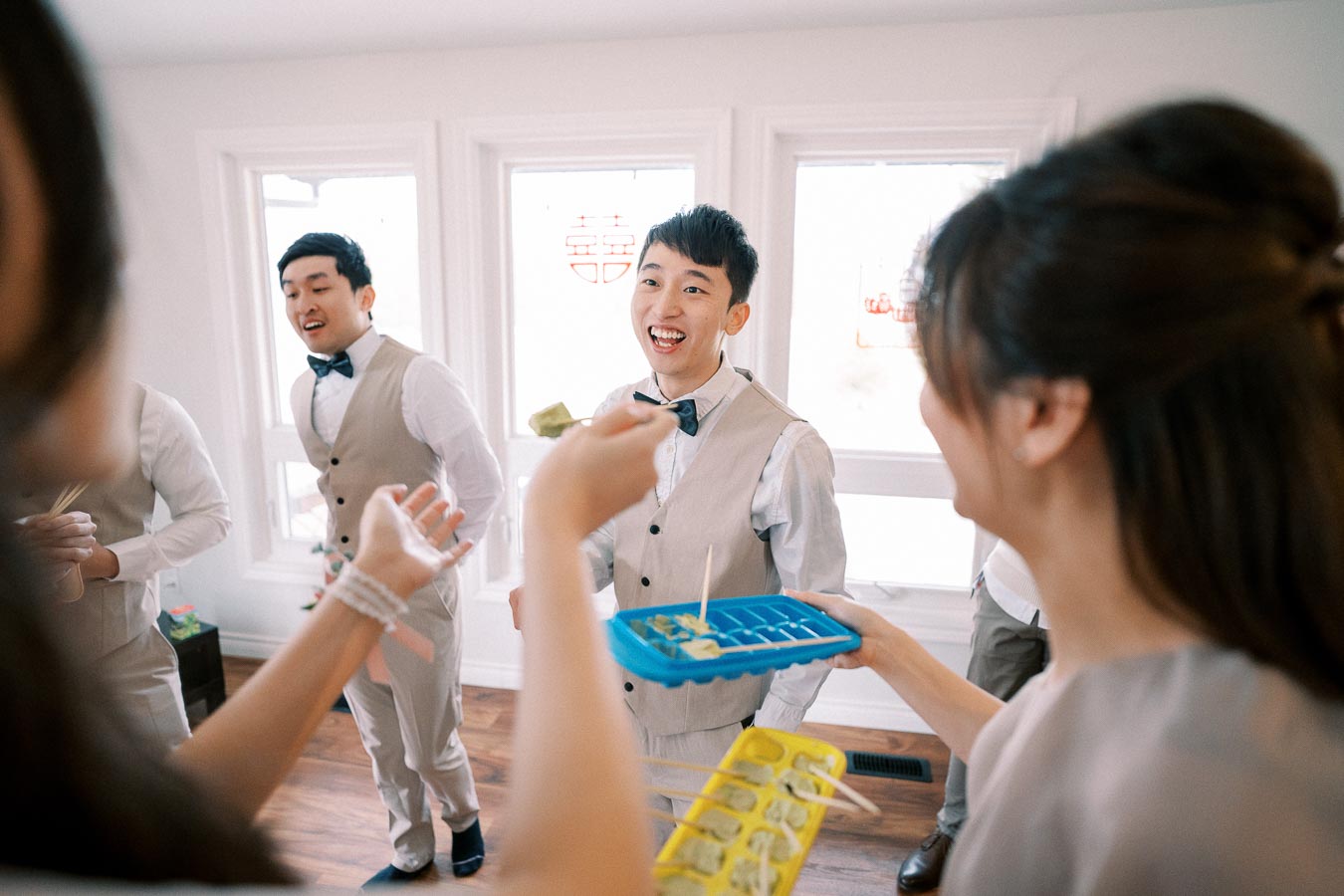 Groomsmen enjoying a lively icebreaker game at a wedding party, wearing matching beige vests and bow ties, with bright natural light from a nearby window.