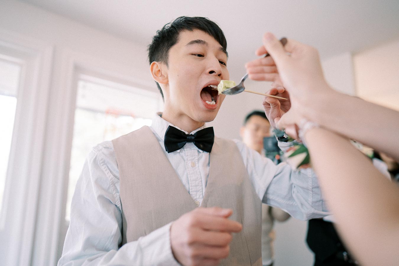 A man wearing a bow tie and vest eagerly takes a bite from a spoon during a joyful indoor gathering.