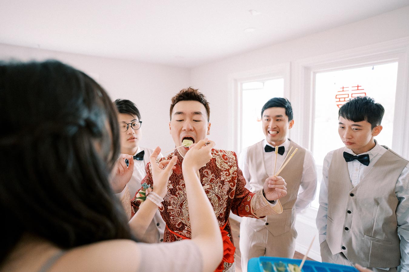 A groom in a traditional embroidered suit participates in a fun wedding game with friends, feeding him food using chopsticks.
