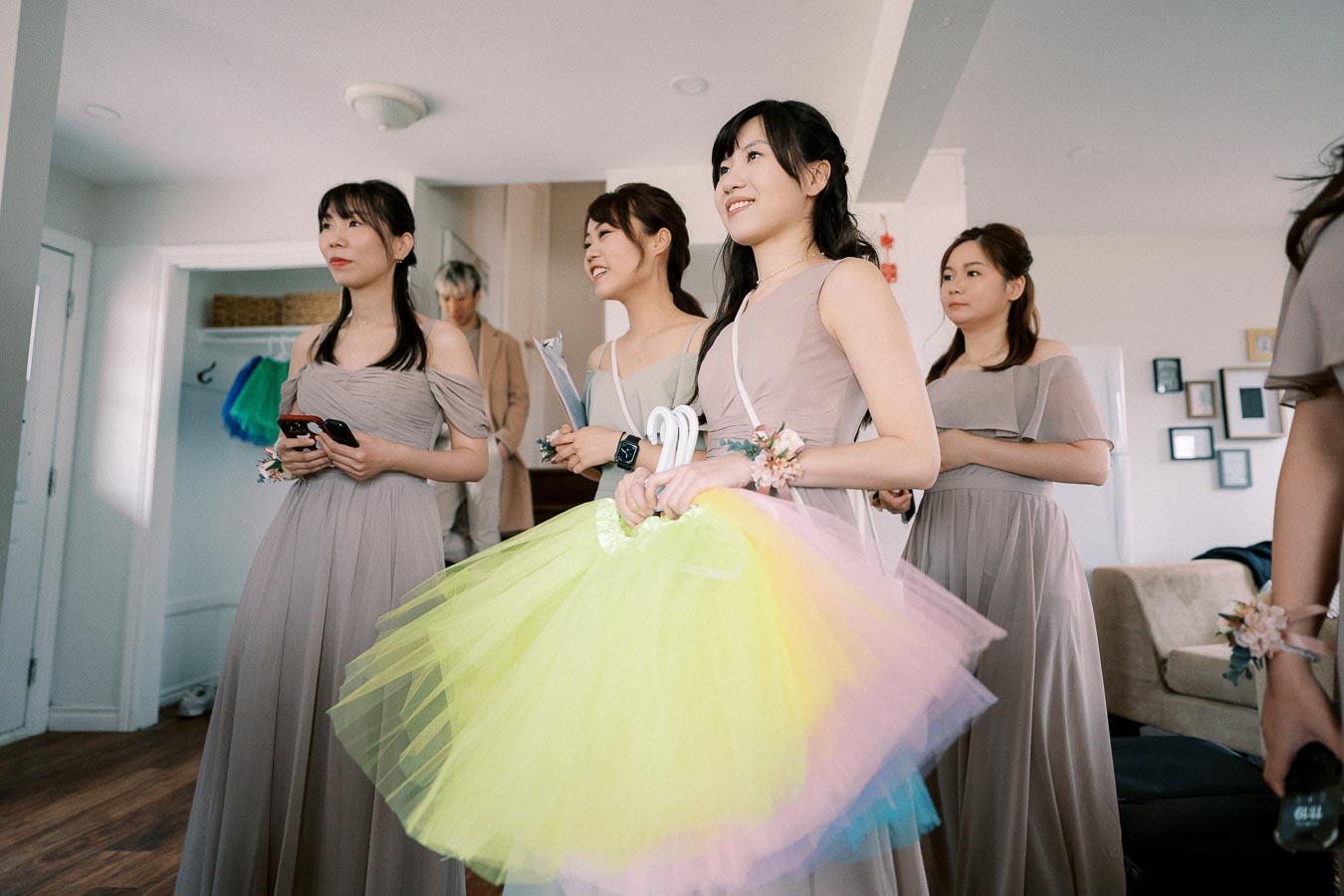 Group of bridesmaids in long dresses, smiling indoors, holding colorful tulle garments, preparing for a wedding ceremony.