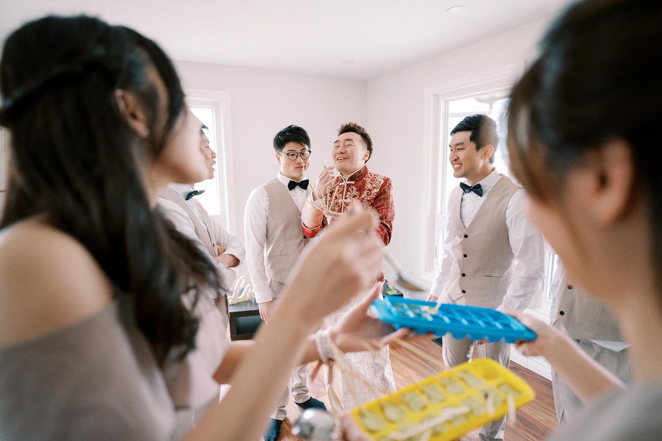 A joyful group of people, including a man in a traditional red embroidered outfit, sharing a moment in a bright room. Some individuals hold trays of ice cubes while others laugh and converse, creating a lively and celebratory atmosphere.