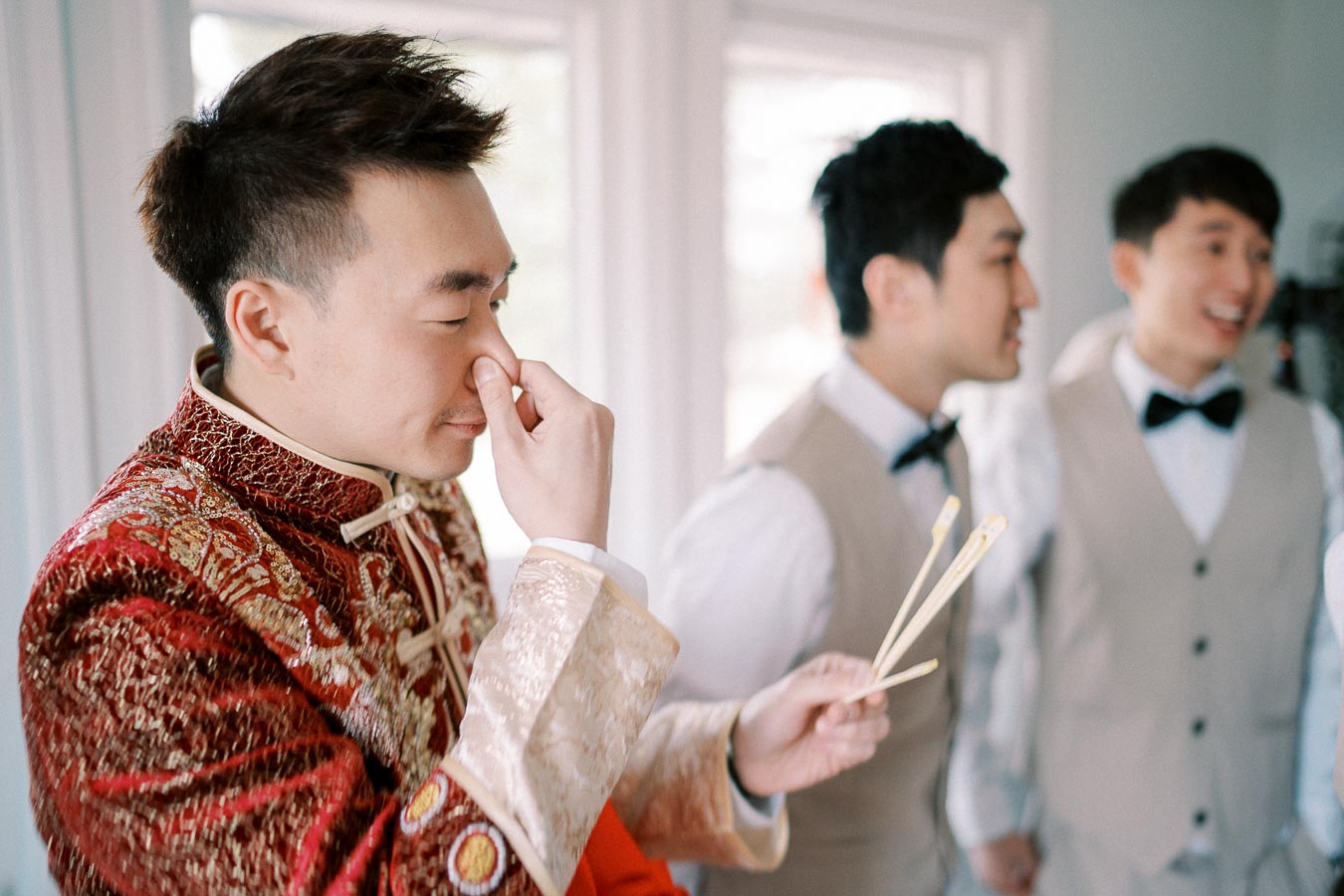 A groom in a traditional red and gold Chinese wedding outfit holds chopsticks and playfully pinches his nose, with groomsmen in the background, preparing for a wedding ceremony.