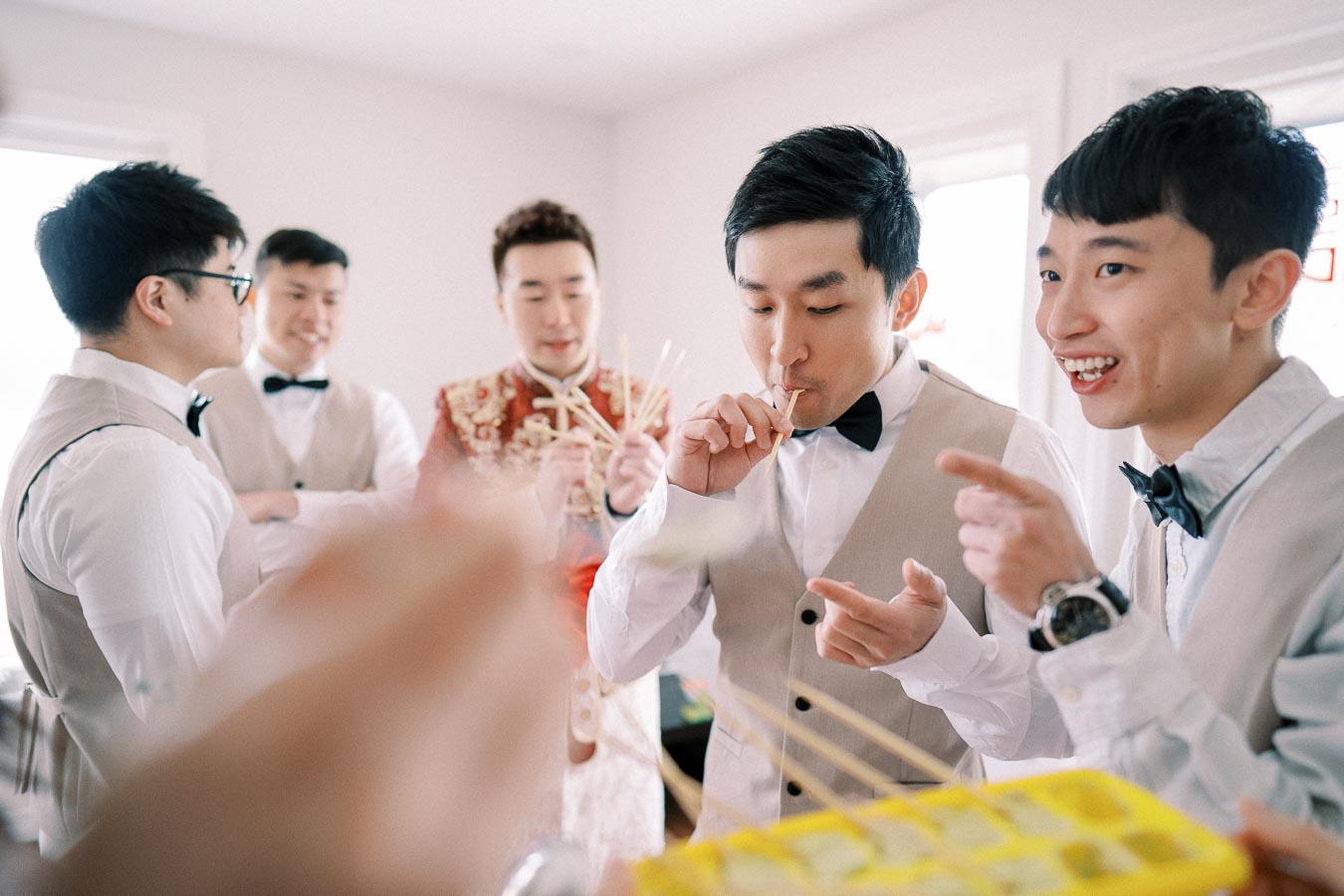 A group of men in formal attire enjoying a game or activity indoors, with one person playfully using a straw from a yellow container. The atmosphere is cheerful and lighthearted.