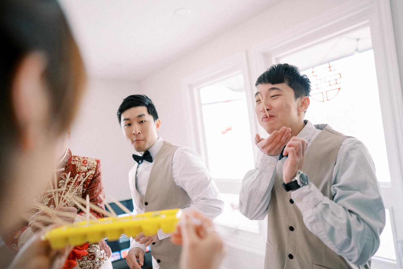 A group of people participating in a playful wedding game involving chopsticks and a yellow ice cube tray, wearing formal attire in a brightly lit room.